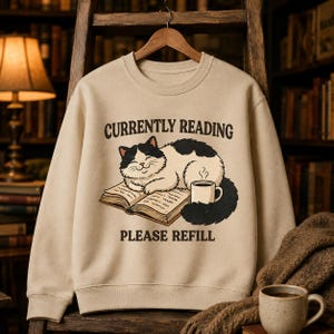 Puede incluir: Una sudadera beige de cuello redondo con el gráfico de un gato blanco y negro tumbado sobre un libro abierto junto a una taza humeante. El texto CURRENTLY READING aparece arriba y PLEASE REFILL abajo en letras de color marrón oscuro.