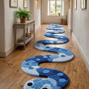 Puede incluir: Una alfombra larga y sinuosa con forma de serpiente que presenta una superficie texturizada en varios tonos de azul, incluyendo azul claro, azul medio y azul oscuro. La alfombra tiene una cabeza redondeada con dos ojos oscuros y una lengua.