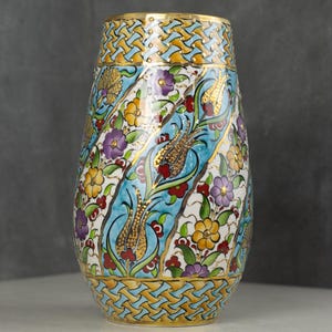 Op de afbeelding: Een decoratieve keramische vaas met een kleurrijk bloemmotief. De vaas heeft een lichtblauwe achtergrond met gouden accenten en een herhalend patroon aan de boven- en onderkant. Het bloemmotief omvat gele, paarse en rode bloemen.