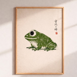 May include: A framed print featuring a green frog in a Japanese woodblock style. The frog has large, round eyes and is set against a cream-colored background. Japanese characters are printed on the right side of the image.