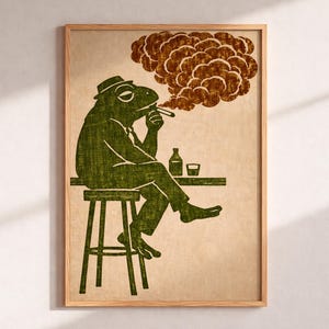 May include: A framed art print features a green frog in a suit and hat, smoking a cigar while sitting at a bar. The frog is depicted with a bottle and a glass on the bar. The background is a beige color, and the smoke is brown.