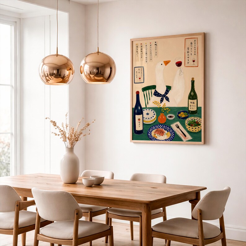 May include: A rectangular wall art print featuring a white goose wearing a blue polka dot scarf, sitting at a green table with wine, plates of food, and Japanese text. The illustration is framed in light wood and displayed on a white wall.