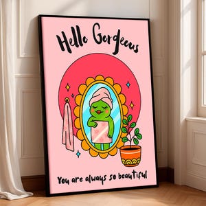 Può includere: Una stampa artistica rosa incorniciata con le parole "Hello Gorgeous" e "You are always so beautiful". L'illustrazione presenta una rana in un asciugamano che si guarda allo specchio. Una pianta in vaso e un asciugamano sono anche nell'immagine.