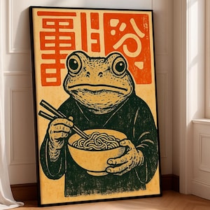 Può includere: Stampa artistica incorniciata raffigurante una rana che mangia ramen con le bacchette. La rana indossa una veste verde scuro e tiene in mano una ciotola di noodles. Lo sfondo è beige chiaro con caratteri giapponesi arancioni.
