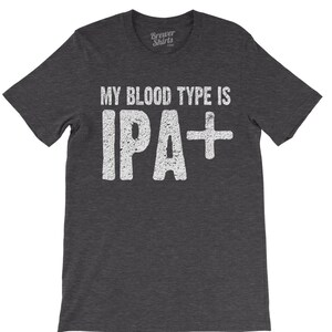 Puede incluir: Camiseta gris oscuro con el texto "MY BLOOD TYPE IS IPA+" en letras blancas desgastadas. La camiseta tiene cuello redondo y mangas cortas. El logotipo de Brewer Shirts est&aacute; en la esquina superior derecha.