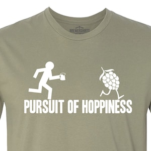 May include: A khaki green t-shirt with a white graphic of a stick figure running with a beer mug and a hop cone running with the text "PURSUIT OF HOPPINESS".