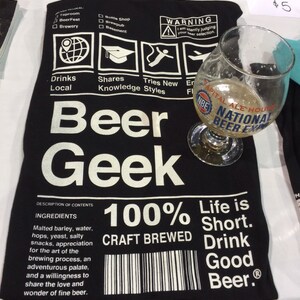Puede incluir: Camiseta negra con texto blanco que dice "Beer Geek" y "100% Craft Brewed". La camiseta tambi&eacute;n tiene una etiqueta de advertencia que dice "Estoy juzgando silenciosamente tu selecci&oacute;n de cerveza."