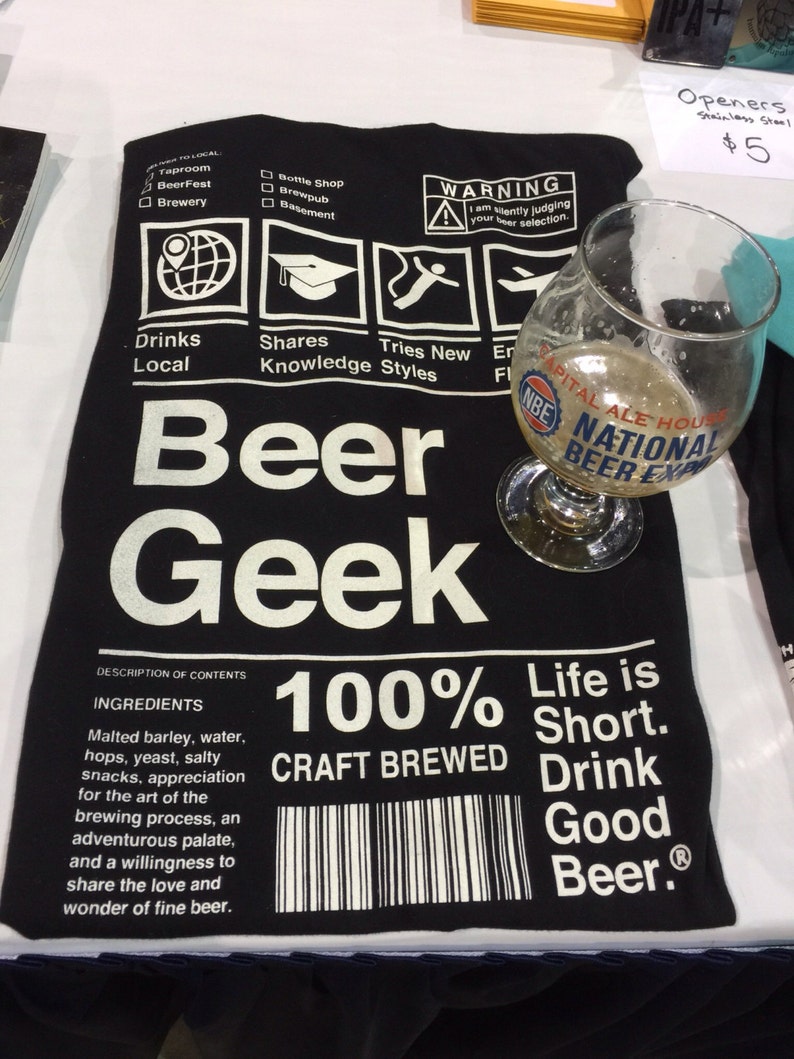Puede incluir: Camiseta negra con texto blanco que dice "Beer Geek" y "100% Craft Brewed". La camiseta tambi&eacute;n tiene una etiqueta de advertencia que dice "Estoy juzgando silenciosamente tu selecci&oacute;n de cerveza."