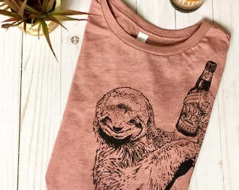 May include: A pink t-shirt with a black and white illustration of a sloth holding a bottle of beer. The beer bottle has the text "Slow Brewed" on it.