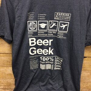 Puede incluir: Una camiseta azul oscuro con un dise&ntilde;o gr&aacute;fico blanco que dice "Beer Geek" e incluye una lista de verificaci&oacute;n de actividades relacionadas con la cerveza. El gr&aacute;fico tambi&eacute;n incluye el texto "100% Craft Brewed" y "Life is Short. Drink Good Beer."