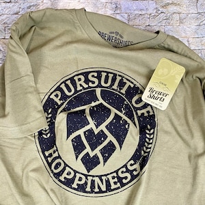 May include: A green t-shirt with a black and white circular graphic that says "Pursuit of Happiness" with a hop leaf inside the circle. The graphic has a distressed look. The t-shirt has a tag that says "Genuine Brewer Shirts Quality Gear Since 2000."