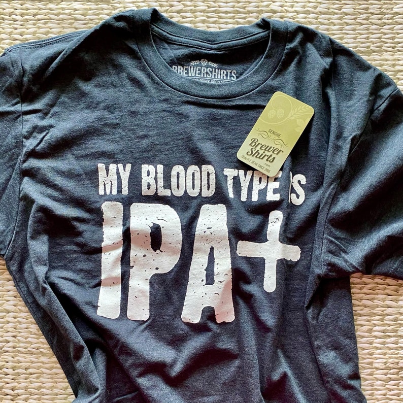 Puede incluir: Camiseta gris oscuro con el texto "MY BLOOD TYPE IS IPA+" en letras blancas. La camiseta tiene una etiqueta que dice "Genuine Brewer Shirts Quality Gear Since 2005". La camiseta est&aacute; hecha de un material suave.