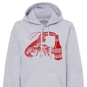 Puede incluir: Una sudadera con capucha gris con una ilustración roja de una langosta sosteniendo una botella de cerveza. La botella de cerveza tiene el texto "MULLER LAGER" en la etiqueta.
