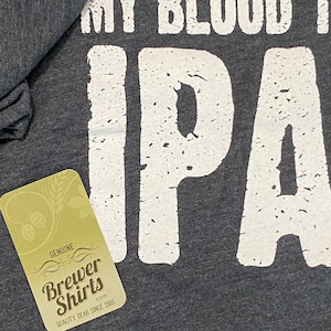 Puede incluir: Camiseta gris oscuro con el texto "MY BLOOD IS IPA" en letras blancas desgastadas. Una etiqueta con el logotipo de "Brewer Shirts" es visible en la esquina inferior izquierda. La camiseta es de calidad.