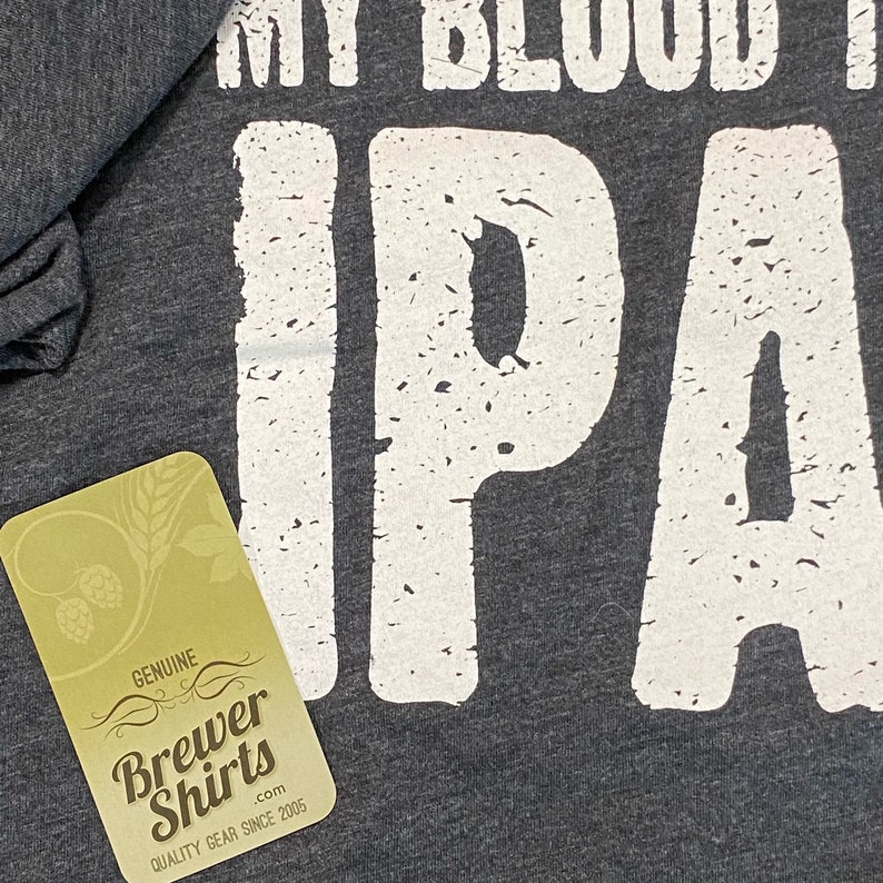 Puede incluir: Camiseta gris oscuro con el texto "MY BLOOD IS IPA" en letras blancas desgastadas. Una etiqueta con el logotipo de "Brewer Shirts" es visible en la esquina inferior izquierda. La camiseta es de calidad.