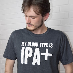 Puede incluir: Camiseta gris oscuro con el texto blanco "MY BLOOD TYPE IS IPA+". La camiseta es de cuello redondo y la lleva una persona. La imagen est&aacute; tomada contra una pared de ladrillos blancos.