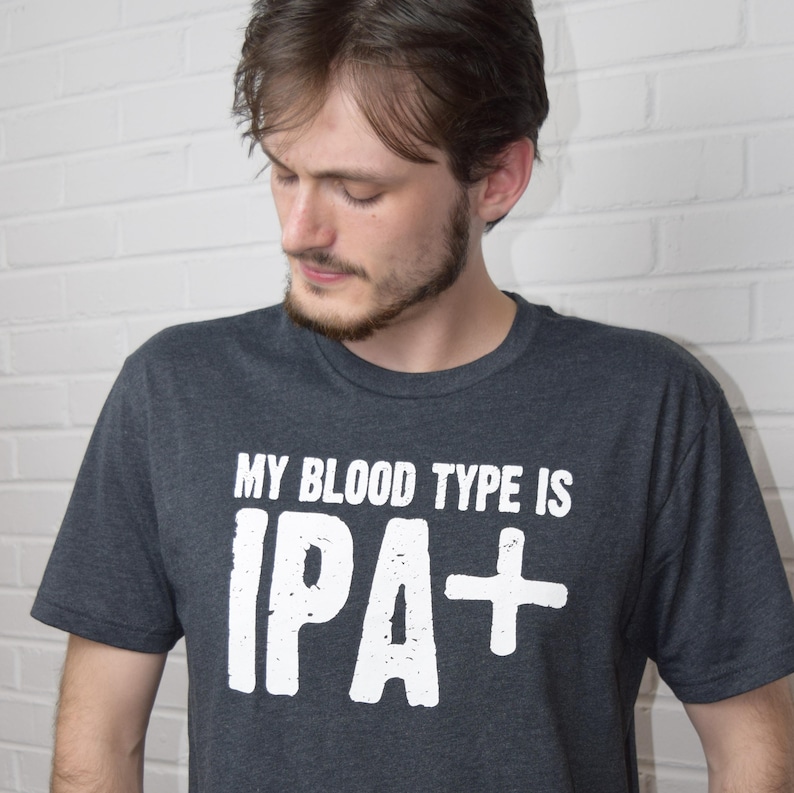 Puede incluir: Camiseta gris oscuro con el texto blanco "MY BLOOD TYPE IS IPA+". La camiseta es de cuello redondo y la lleva una persona. La imagen est&aacute; tomada contra una pared de ladrillos blancos.