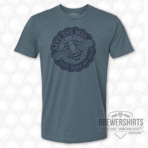 Puede incluir: Una camiseta azul verdosa con un diseño circular que presenta una abeja y el texto "SAVE THE BEES THEY MAKE MY MEAD." La camiseta también tiene el texto "BREWERSHIRTS" y "LIFE IS SHORT. DRINK GOOD BEER."