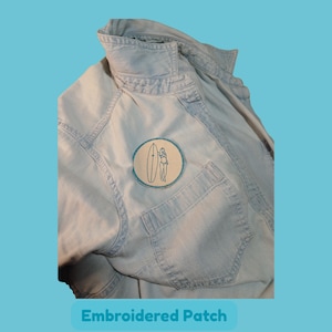 Peut inclure: Une chemise en jean bleu clair avec un écusson brodé circulaire sur la poche poitrine. L'écusson présente un fond crème avec une illustration au trait bleu foncé d'une personne debout à côté d'une planche de surf, bordée de bleu.