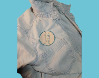 Puede incluir: Una camisa de mezclilla azul claro con un parche bordado circular en el bolsillo del pecho. El parche tiene un fondo color crema con una ilustración de contorno azul oscuro de una persona junto a una tabla de surf, con un borde azul.