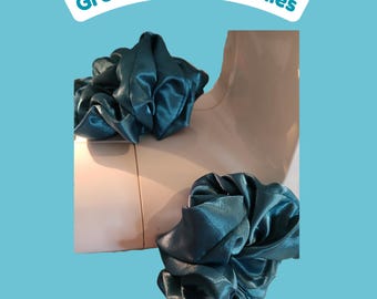Puede incluir: Dos coleteros de color verde azulado oscuro con un acabado satinado liso, mostrados sobre un maniquí de color claro. El texto "Green Blue Scrunchies" aparece impreso en una banda blanca en la parte superior sobre un fondo azul claro.