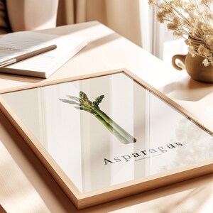 May include: A framed print featuring a vibrant green asparagus spear against a neutral background. The word "Asparagus" is printed in a stylish font below the vegetable. The frame is made of light-colored wood.