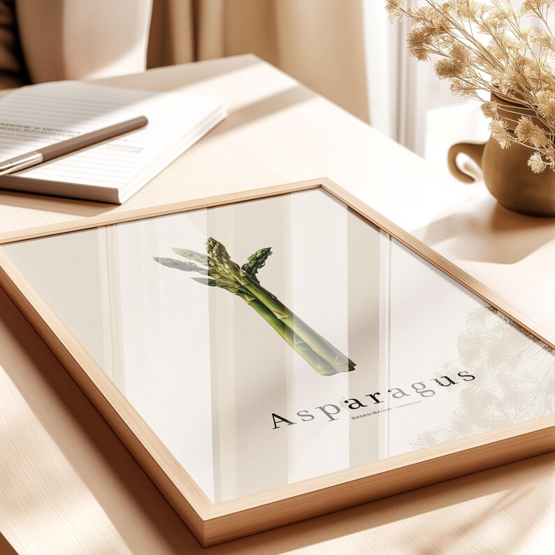 May include: A framed print featuring a vibrant green asparagus spear against a neutral background. The word "Asparagus" is printed in a stylish font below the vegetable. The frame is made of light-colored wood.