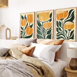 Può includere: Tre stampe artistiche floreali incorniciate. Ogni stampa presenta un design floreale arancione stilizzato con foglie verdi su sfondo crema. Le cornici sono di colore legno chiaro. Le stampe sono esposte sopra un letto con cuscini e coperte.