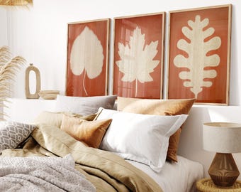 May include: Three framed prints featuring cream-coloured leaf designs on a reddish-orange backdrop. The artwork hangs above a bed with neutral-toned bedding and pillows, creating a warm bedroom setting. The frames are made of wood.