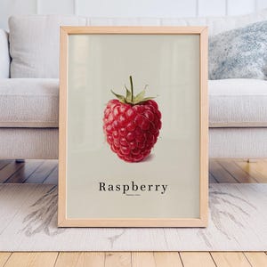 May include: A framed print featuring a detailed illustration of a ripe red raspberry. The fruit is centered against a light background, with the word "Raspberry" printed below. The frame is made of light-colored wood.