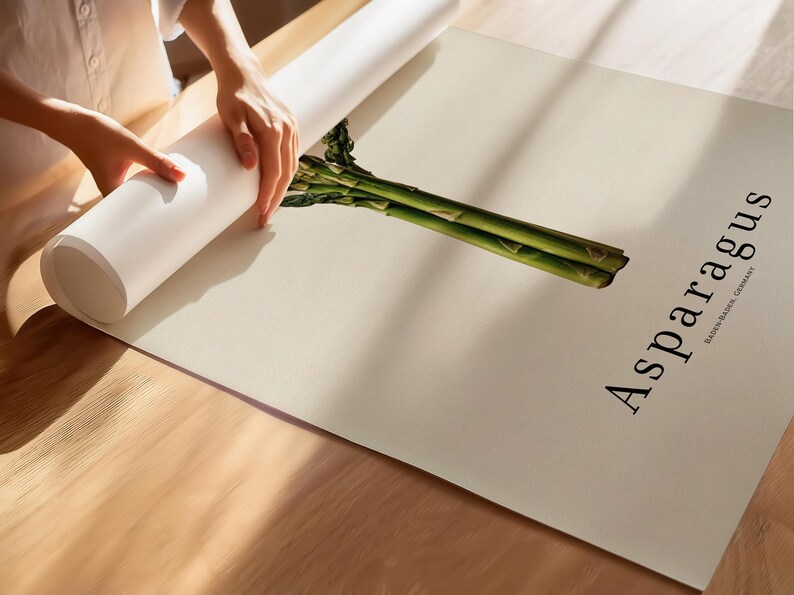 May include: A rolled white poster with the word "Asparagus" in black font, with a green asparagus illustration. The poster is being unrolled on a light beige surface. The text "BADEN-BADEN, GERMANY" is below the word "Asparagus".