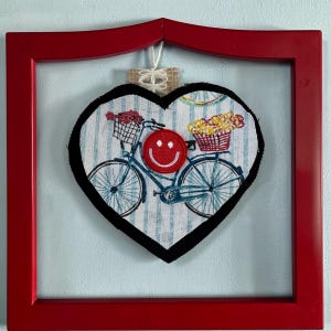 Puede incluir: Un adorno de pared en forma de corazón con un marco rojo. La obra de arte presenta una bicicleta azul con cestas de flores y una cara sonriente roja. El fondo tiene rayas azules y blancas. Una pequeña etiqueta de arpillera cuelga de la parte superior.