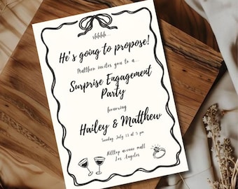 Peut inclure: Une invitation crème avec un texte noir annonçant une fête de fiançailles surprise pour Hailey et Matthew. Le texte comprend "Il va demander en mariage !" et les détails de l'événement. La carte est posée sur une surface en bois.