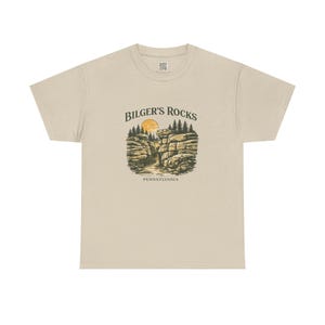 Puede incluir: Camiseta beige con un gráfico de Bilger's Rocks en Pensilvania. El diseño incluye un sol, árboles y formaciones rocosas. El texto "BILGER'S ROCKS" está arqueado sobre la imagen, con "PENNSYLVANIA" debajo.