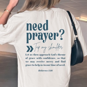Puede incluir: Camiseta color crema con el texto "need prayer? Tap my shoulder" en color turquesa. Debajo, una cita de Hebreos 4:16. La camiseta es de estilo casual.