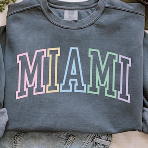 Puede incluir: Una sudadera gris oscuro con la palabra "MIAMI" impresa en la parte delantera en un diseño de bloques de color pastel. Las letras están delineadas en un color más oscuro y la sudadera tiene cuello redondo.