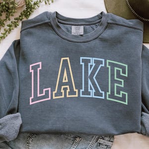 Pode incluir: Um moletom cinza escuro com a palavra "LAKE" em letras grandes e coloridas. As letras s&atilde;o contornadas em cores pastel: rosa, amarelo, azul e verde. O moletom &eacute; feito de um material macio e tem gola redonda.