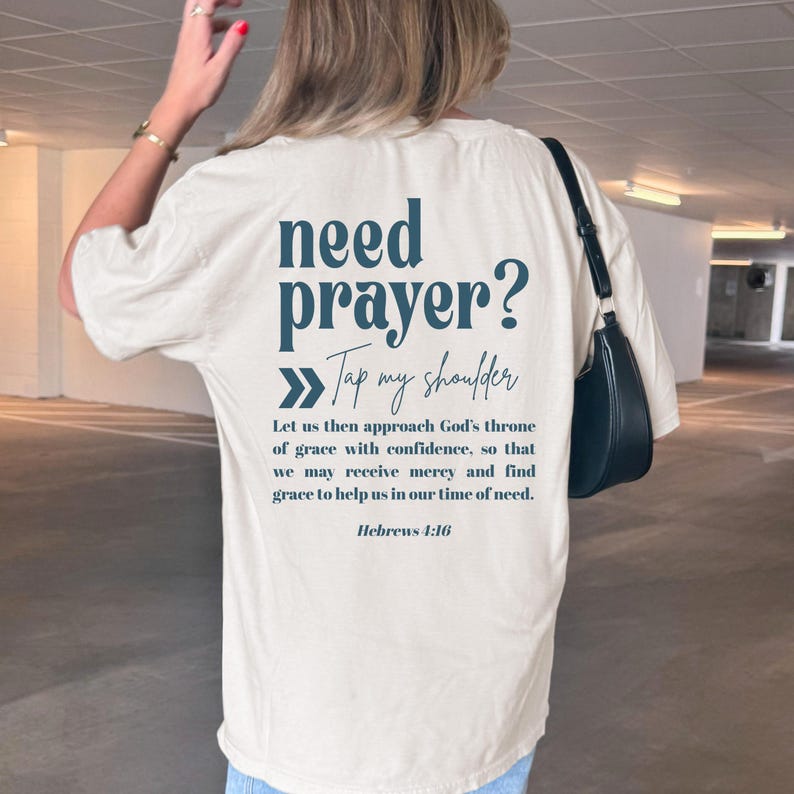 Puede incluir: Camiseta color crema con el texto "need prayer? Tap my shoulder" en color turquesa. Debajo, una cita de Hebreos 4:16. La camiseta es de estilo casual.