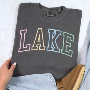Pode incluir: Camisola cinza escuro com a palavra "LAKE" em cores pastel. As letras s&atilde;o delineadas em uma cor mais escura. A camisola est&aacute; dobrada sobre uma superf&iacute;cie branca.