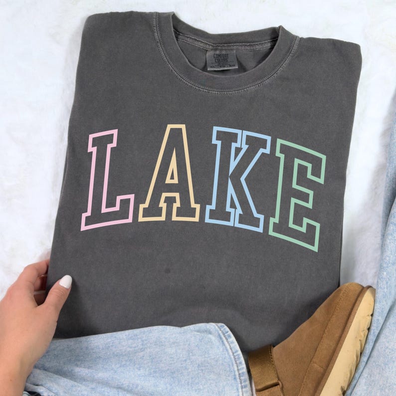 Pode incluir: Camisola cinza escuro com a palavra "LAKE" em cores pastel. As letras s&atilde;o delineadas em uma cor mais escura. A camisola est&aacute; dobrada sobre uma superf&iacute;cie branca.