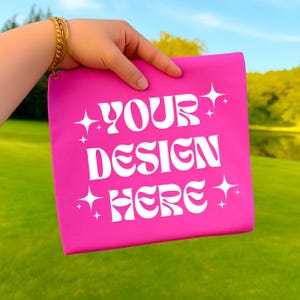 May include: A bright pink square fabric with the text "YOUR DESIGN HERE" in white, surrounded by starburst graphics. The fabric is held up against a backdrop of green grass, a lake, and a blue sky.