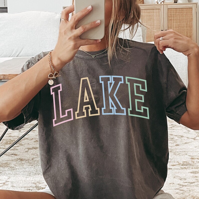 Pode incluir: Camiseta cinza escuro com a palavra "LAKE" em letras grandes. As letras s&atilde;o contornadas em branco com cores pastel no interior, incluindo rosa, amarelo e azul. A camiseta tem um visual vintage e desgastado.