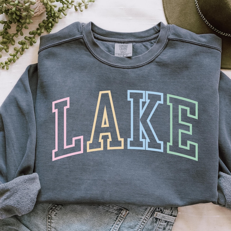 Pode incluir: Um moletom cinza escuro com a palavra "LAKE" em letras grandes e coloridas. As letras s&atilde;o contornadas em cores pastel: rosa, amarelo, azul e verde. O moletom &eacute; feito de um material macio e tem gola redonda.