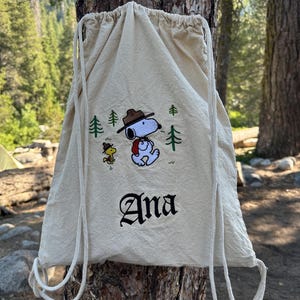 May include: A beige drawstring backpack featuring an embroidered Snoopy and Woodstock design with pine trees. The name "Ana" is embroidered in a gothic font below the cartoon characters. The bag has white drawstrings.