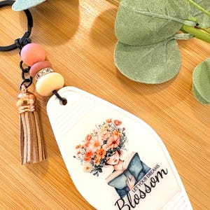 May include: White motel-style keychain with a floral illustration of a woman reading a book, with the text "LET YOUR DREAMS Blossom". The keychain has a black ring, pink and cream beads, a gold tassel, and a black cord.