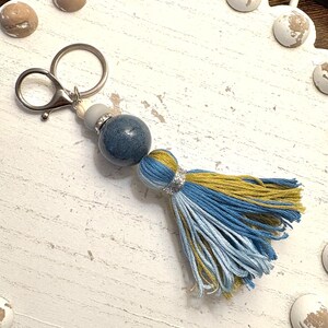 May include: A handmade keychain with a blue and yellow tassel. The keychain features a silver clasp, a gray bead, a blue marbled bead, and a tassel with blue and yellow threads. The background is a white, scalloped tray with decorative globe-shaped accents.
