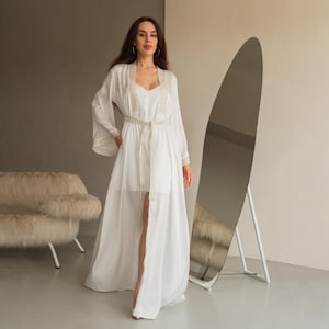 May include: A white, flowing robe with a matching slip dress. The robe features long sleeves with lace trim and a belted waist. A full-length mirror stands to the right, reflecting the room. The overall aesthetic is elegant and airy.