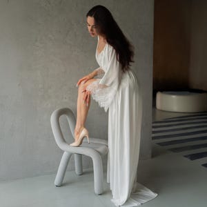 Könnte beinhalten: Ein weißer Satin-Bademantel mit Spitzenbesatz an den Ärmeln und einem passenden Slip. Die Person trägt High Heels und lehnt sich an einen grauen Stuhl.