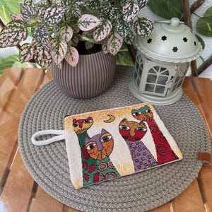 May include: A rectangular, off-white pouch featuring a vibrant cat design. The cats are depicted in various colours and patterns, including red, green, and purple. A small white loop is attached. The pouch rests on a grey woven mat.