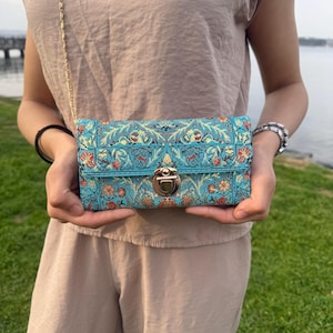 May include: A rectangular clutch with a teal background and a floral pattern in red, orange, and yellow. The clutch has a gold chain strap and a gold clasp. The person is wearing a tan top and pants.